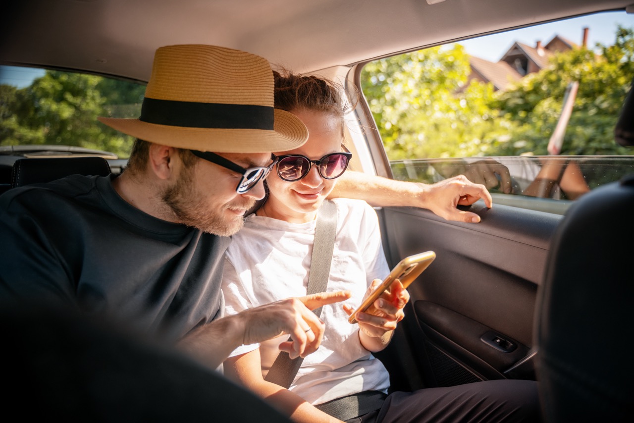 Young cheerful happy couple of travellers sitting in car looking at smartphone screen, travel comfort safety taxi concept